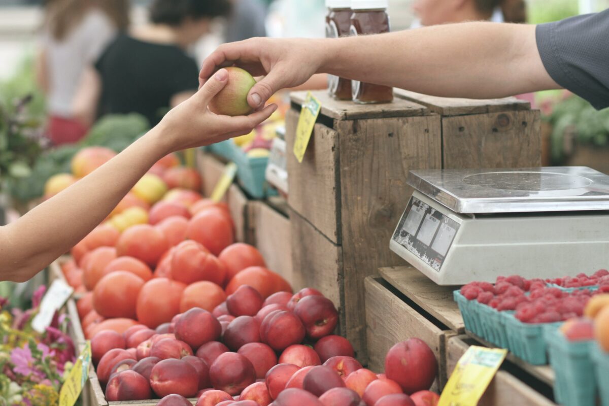 An einem Marktstand übergibt eine verkaufende Person einer kaufenden Person einen Apfel.