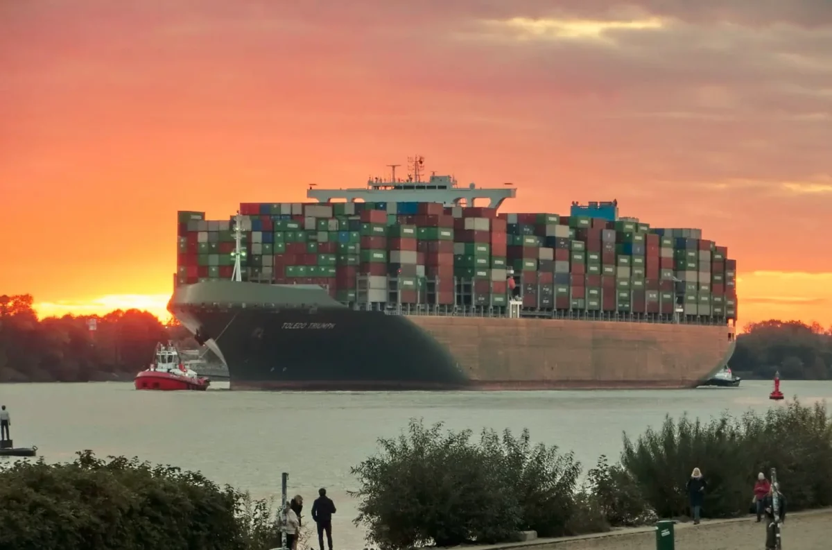 das bild zeigt ein kontainerschiff auf einem fluss im sonnenuntergang. im vordergrund ist eine uferpromenade mit spaziergängern zu sehen.